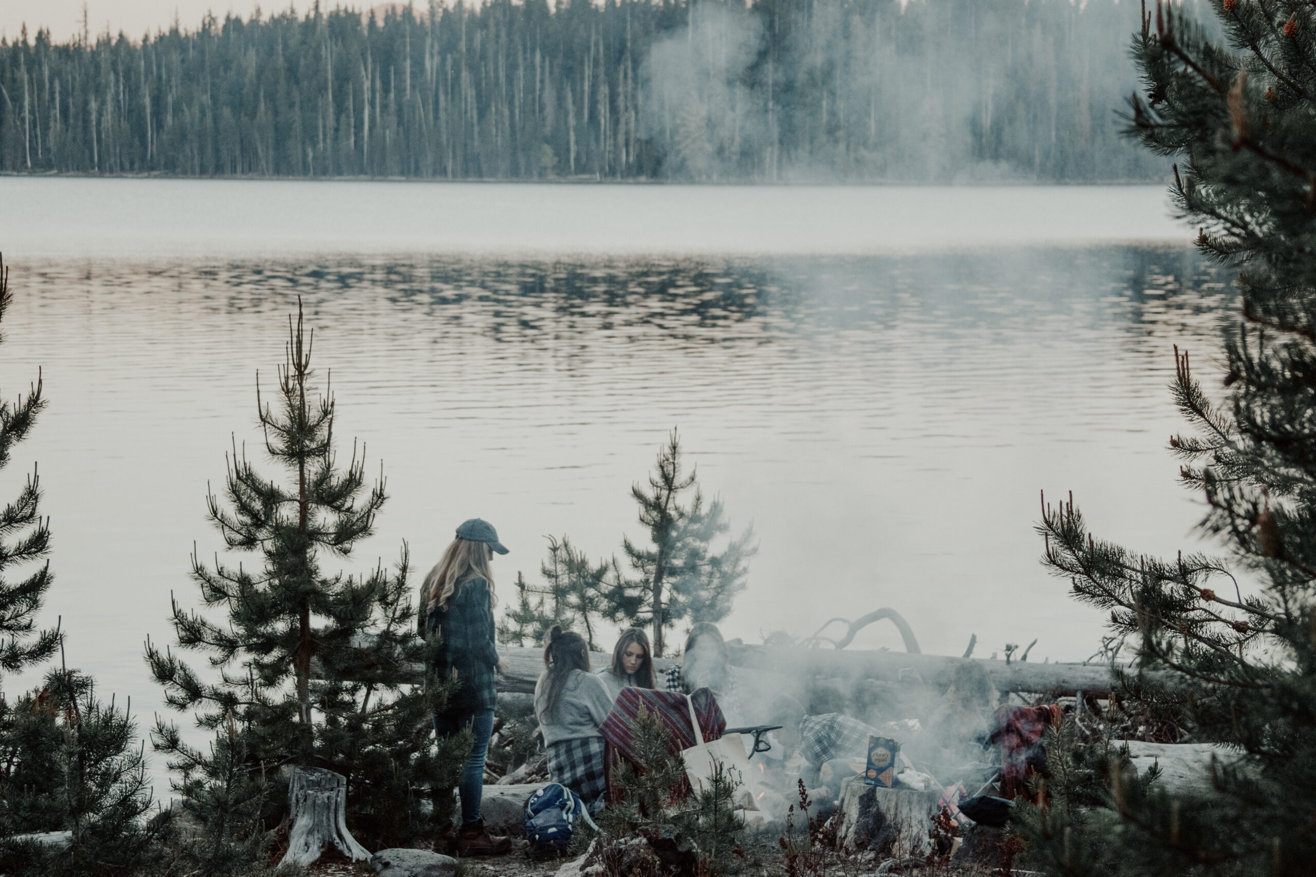 Women stand and sit near a campfire with the lake in the background.