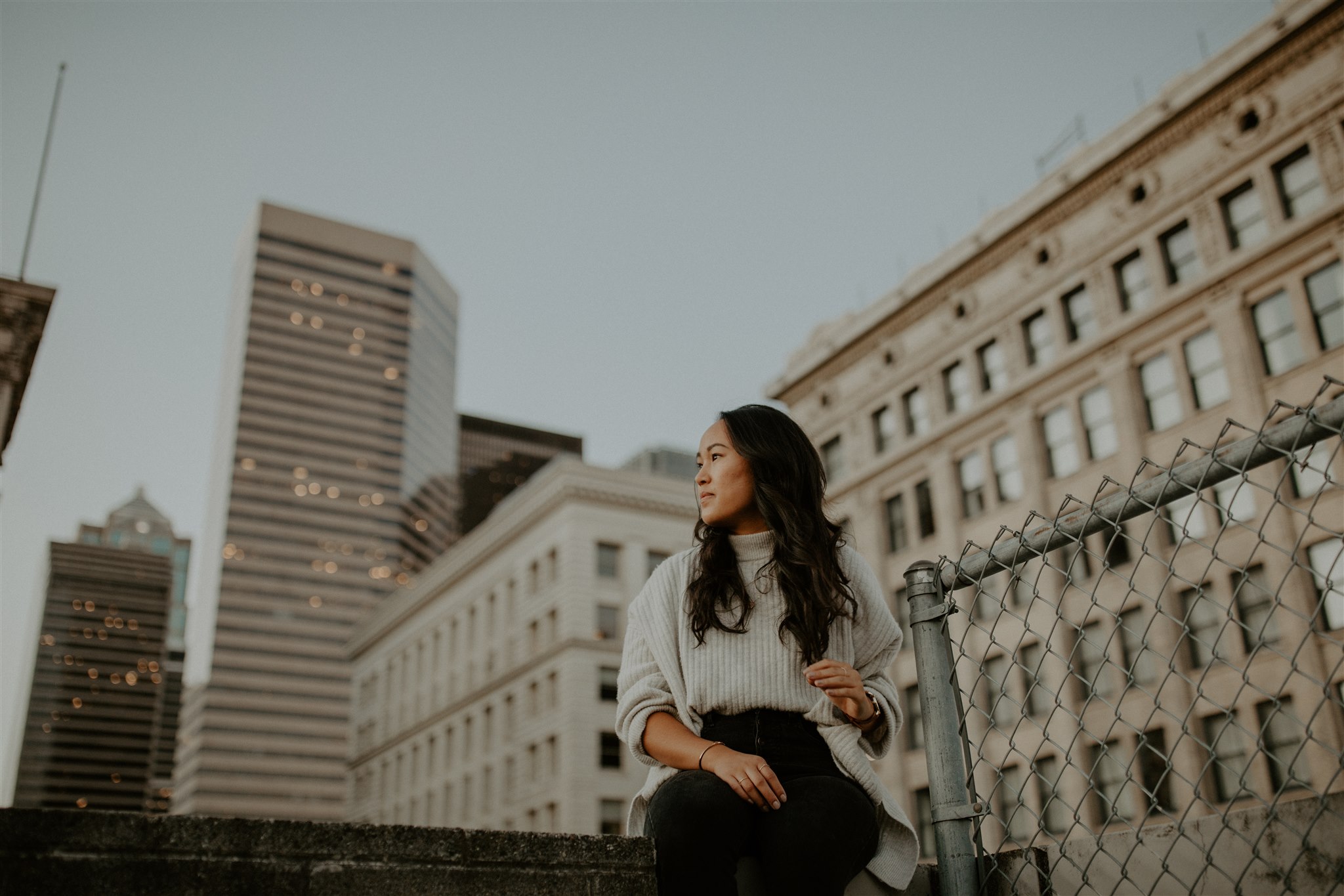 A woman sits on a rooftop with the cityscape behind her.