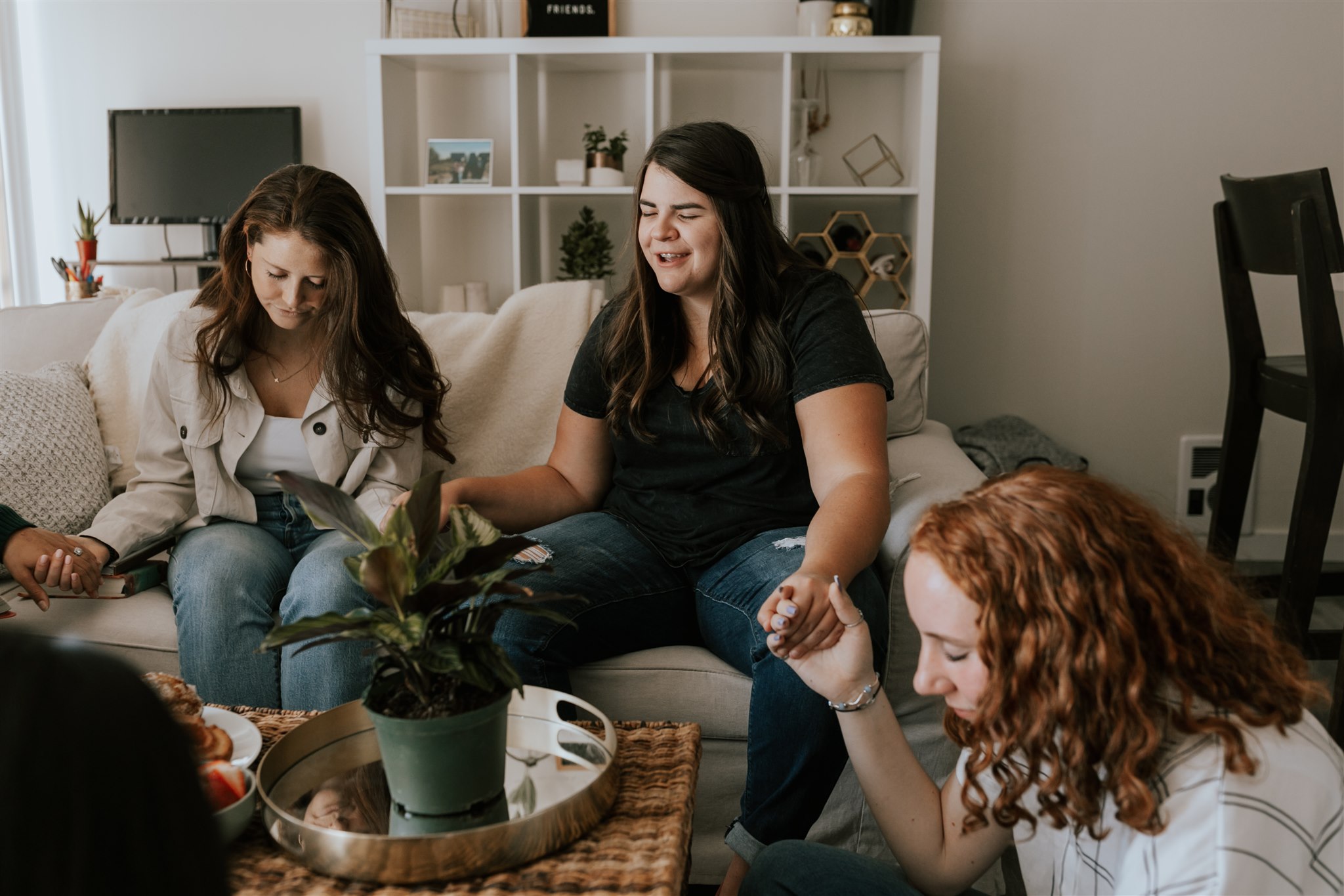 Women gather in a living room to pray for each other, hands joined.