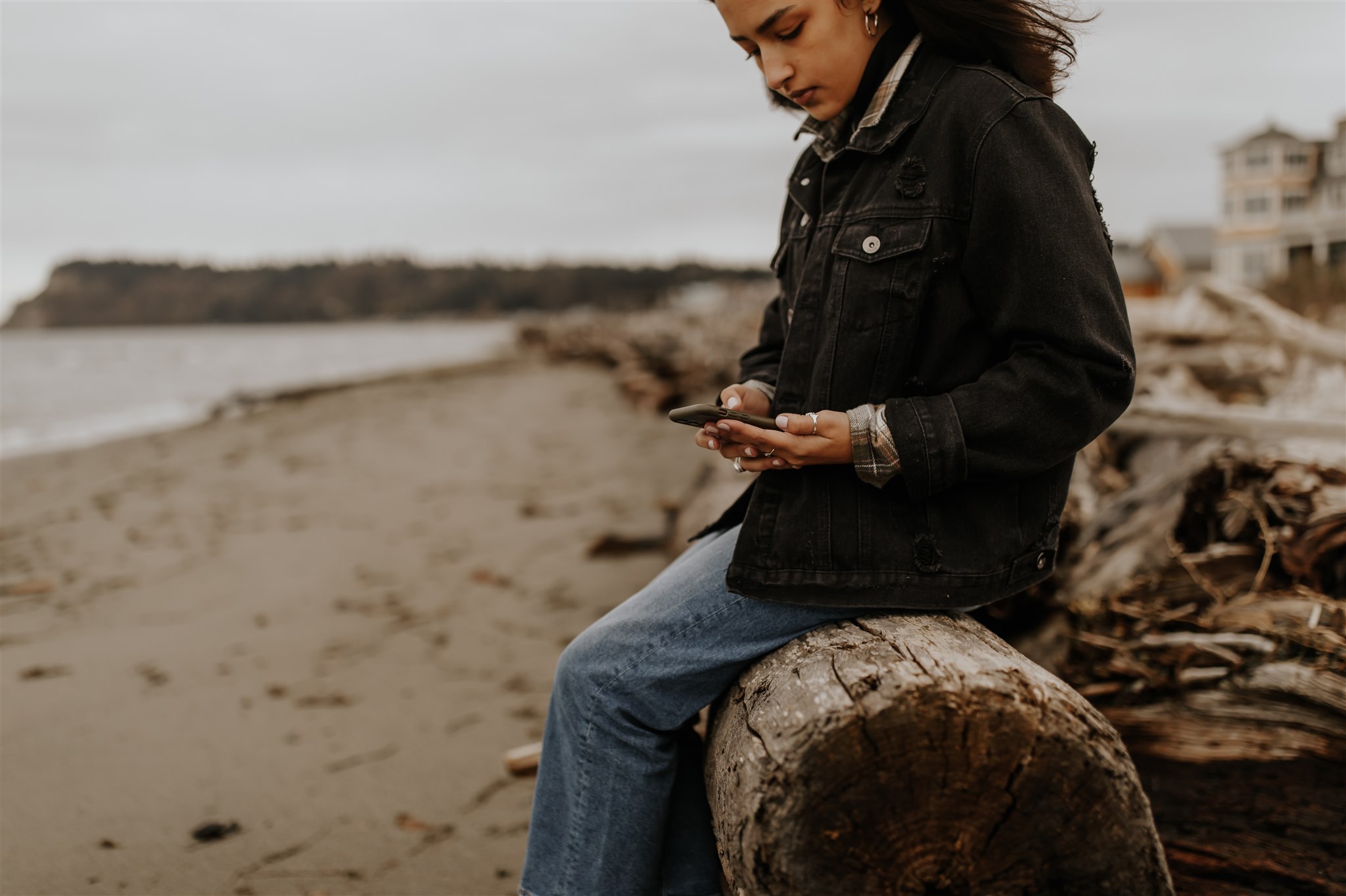 A woman looks down at her phone while she sits on the beach.