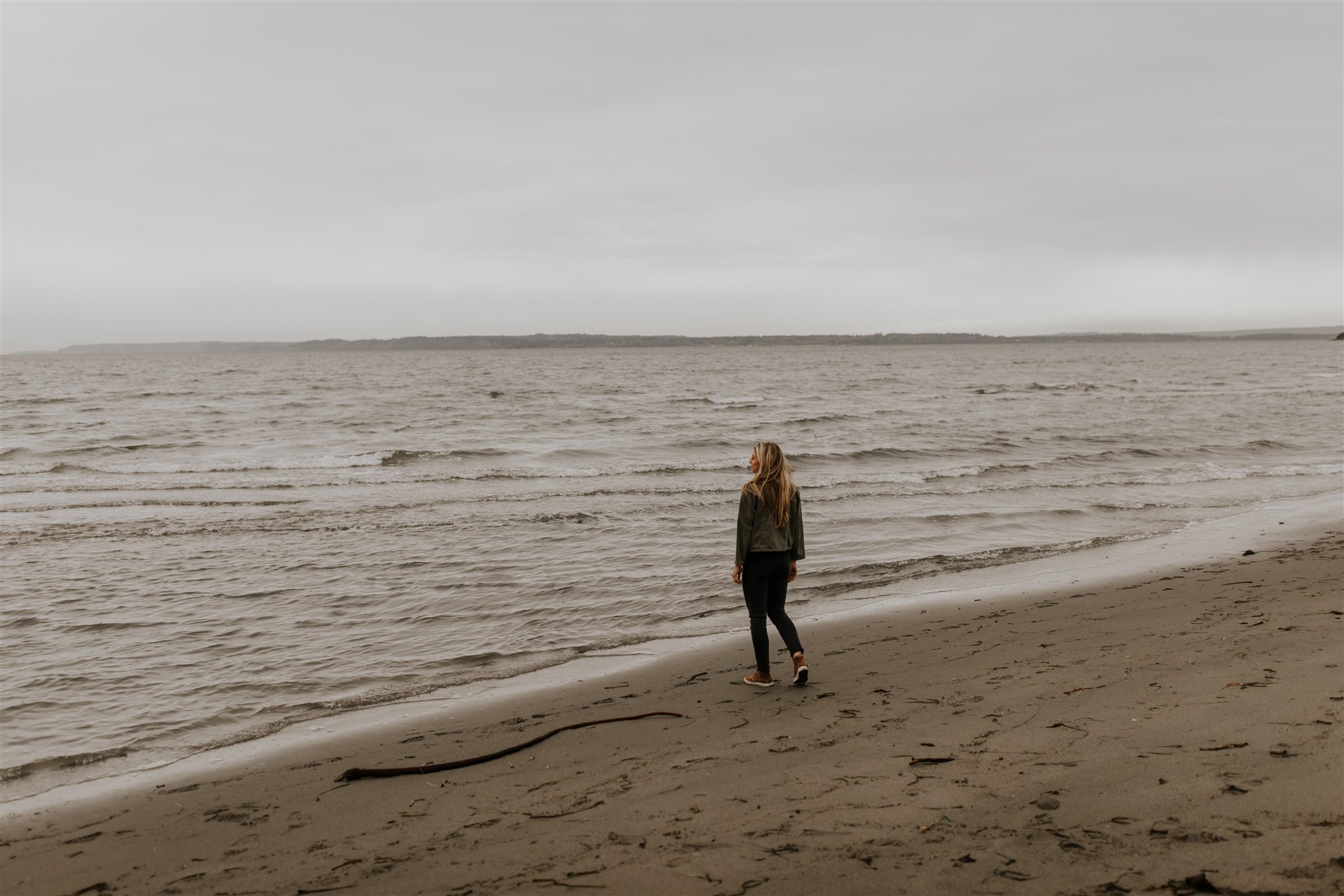 A young woman walks on the beach, bundled in a light jacket.