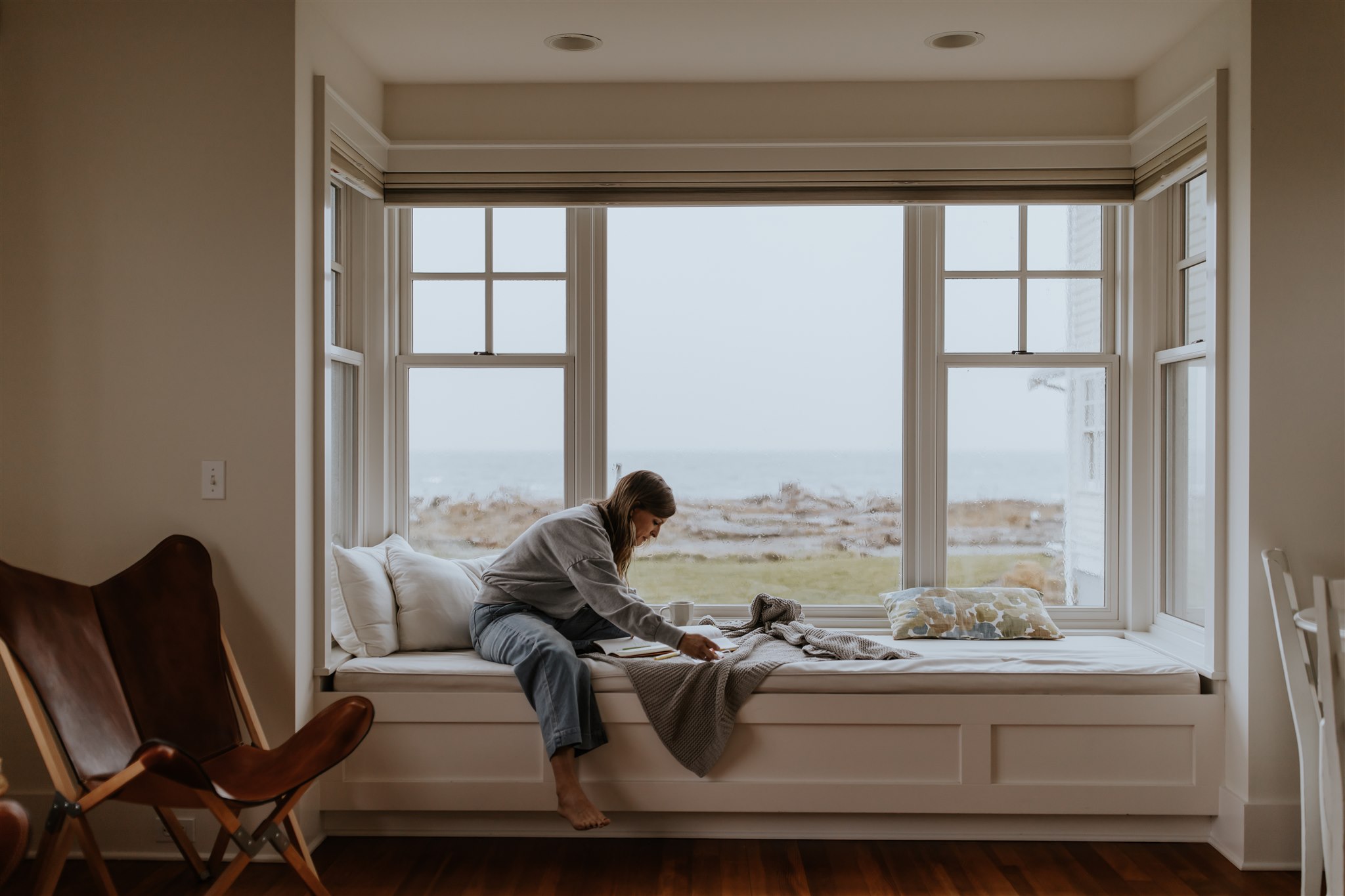 A woman sits on a window seat, the ocean visible from the window, reading her Bible.