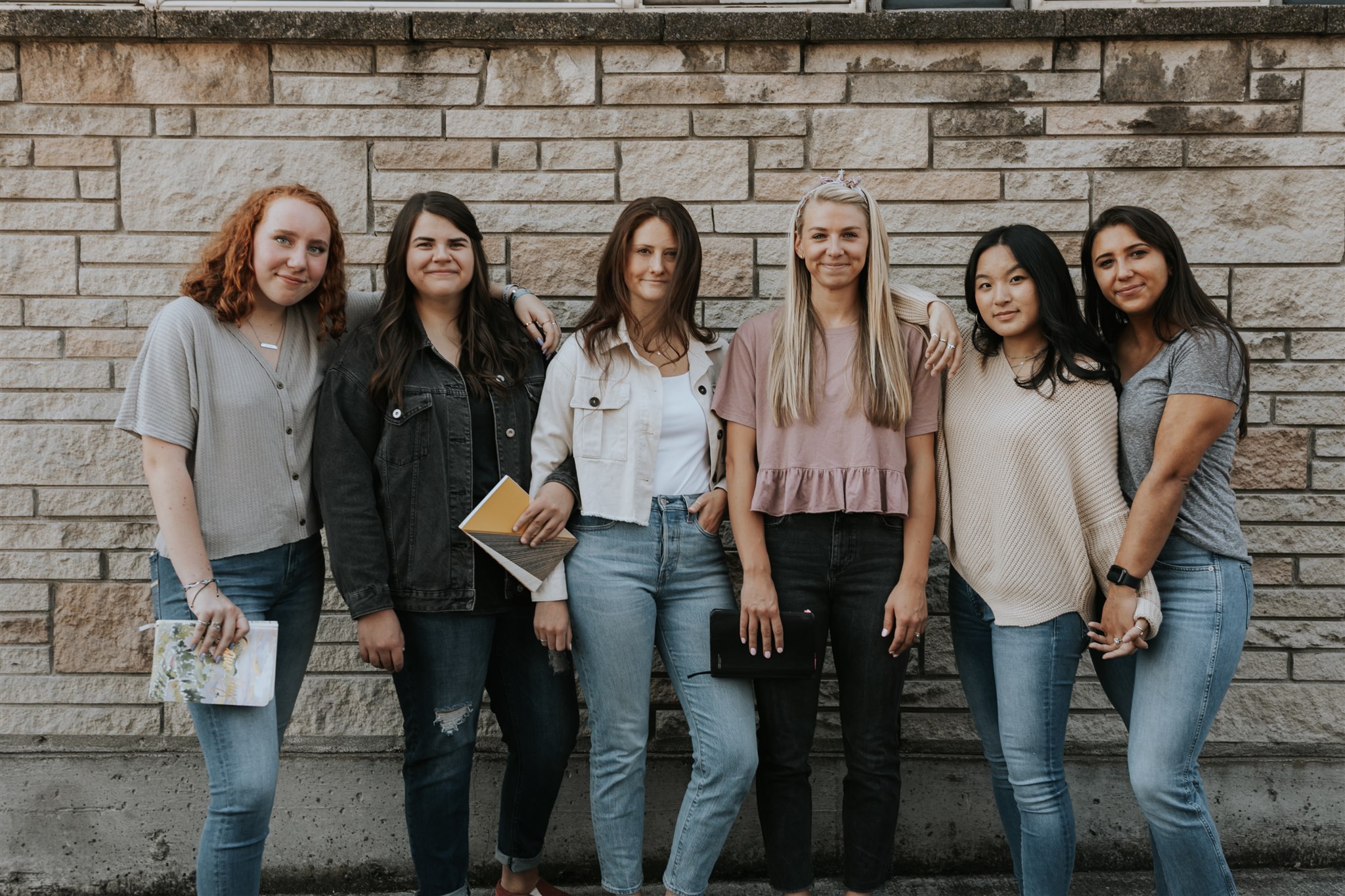 Women stand together outside, smiling at each other.