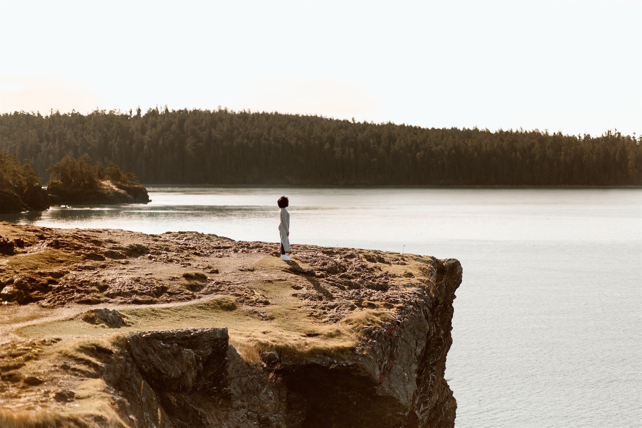 A woman stands at the precipice of a cliff, looking out over the ocean.